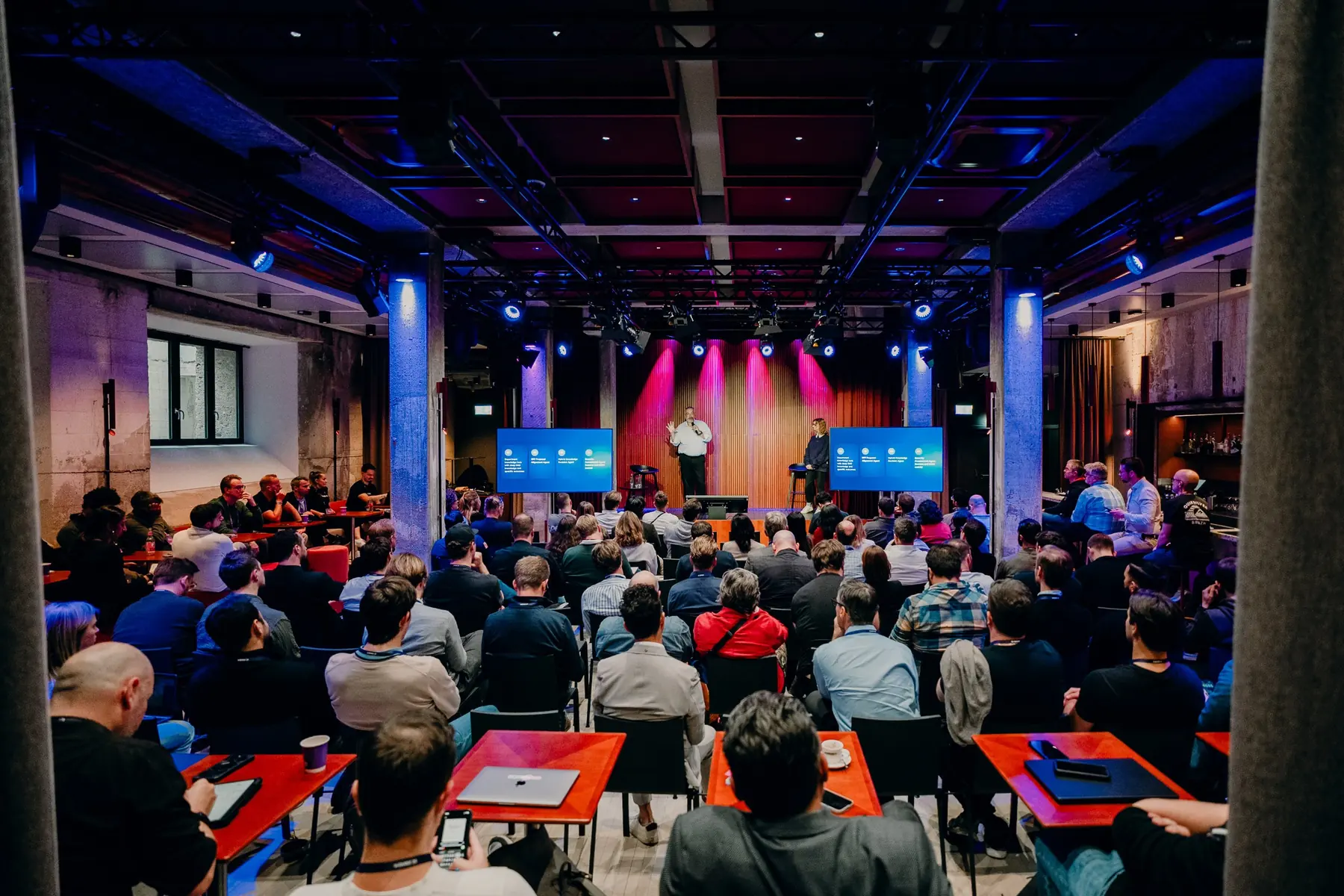 Guests seated in front of a stage with two screens during a talk at a Make event